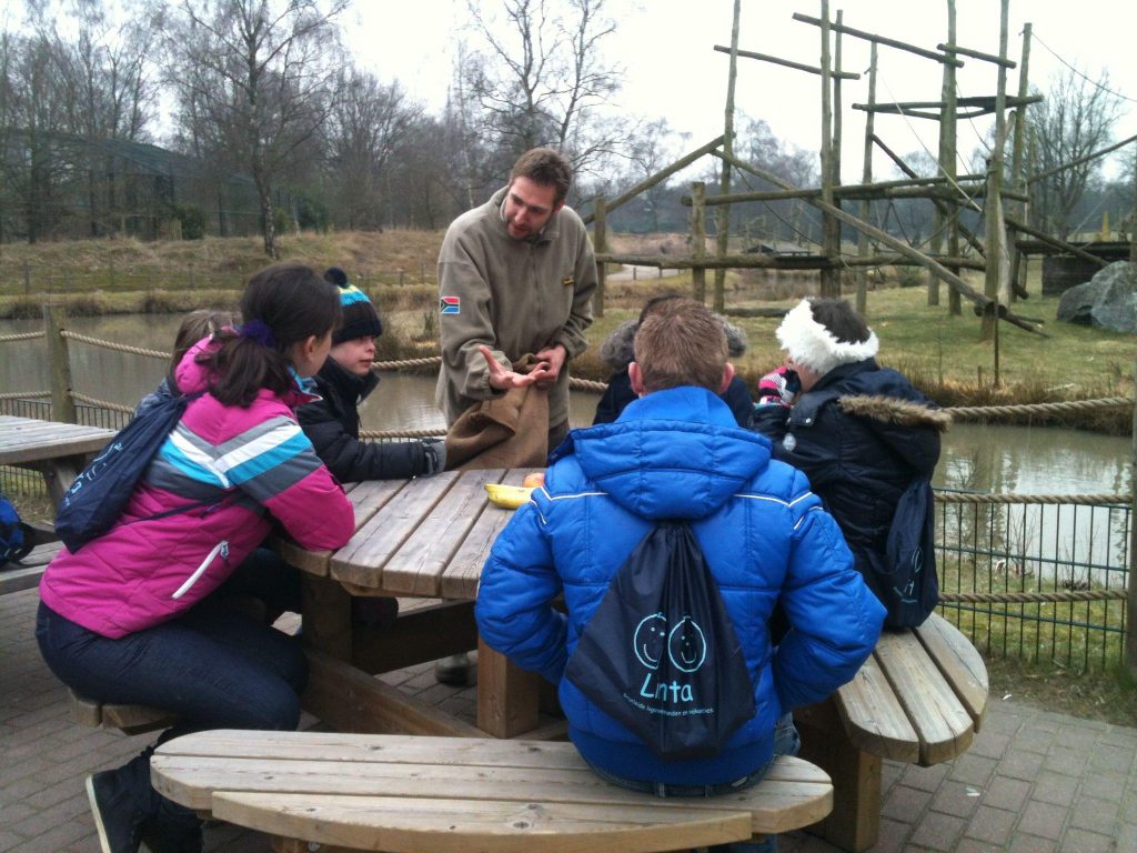 Groepje mensen aan picknicktafel krijgt uitleg over apen in dierentuin.