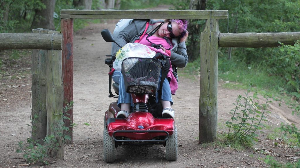 Vrouw rijdt in scootmobiel lachend onder een paaltje in het bos door.